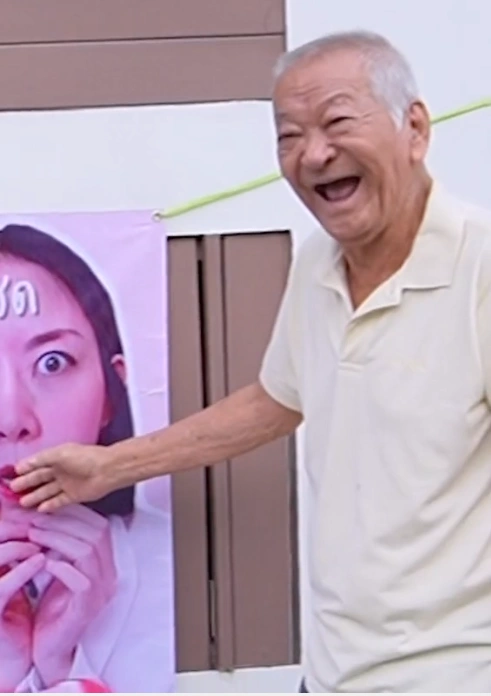 The Thai grandfather holding a phone and interacting with the public while standing next to the matchmaking banner.