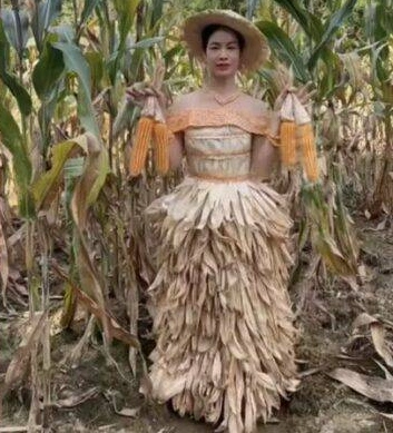 A woman posing in a large, voluminous ballgown made from dried corn husks in a rural village setting.